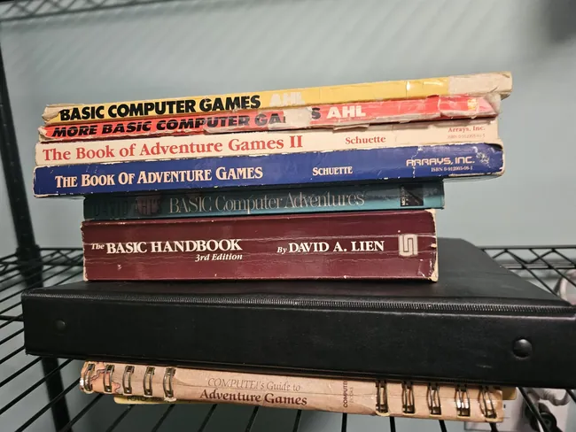 A stack of worn programming books on a wire shelf, three of them by David Ahl: BASIC Computer Games, More BASIC Computer Games and BASIC Computer Adventures.